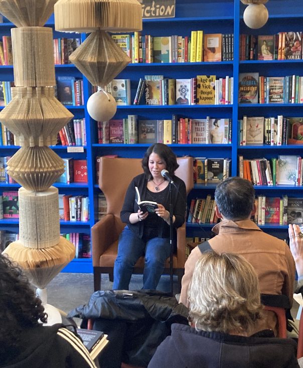 Katherine Quevedo reading at Claw Machine anthology book launch in Up Up Books with a blue bookshelf behind her.