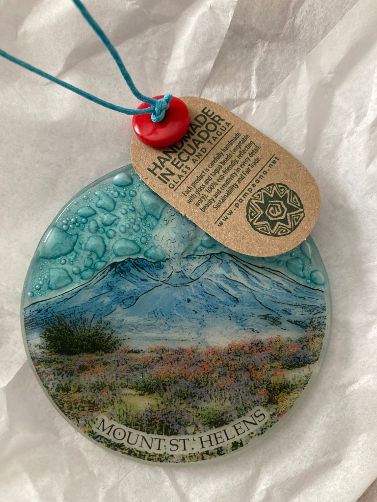 Blue circular glass ornament with a drawing of Mount St. Helens erupting, with bubbles in the glass for the sky, and a paper tag saying "Handmade in Ecuador."