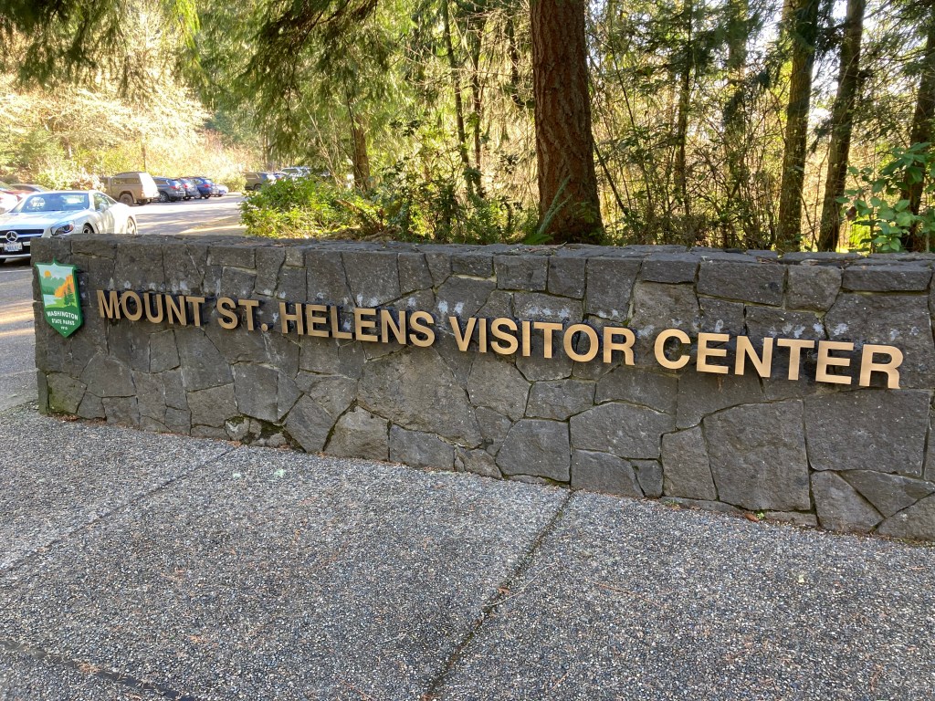 Stone sign with bronze letters saying "Mount St. Helens Visitor Center" with trees and a parking lot in the background.